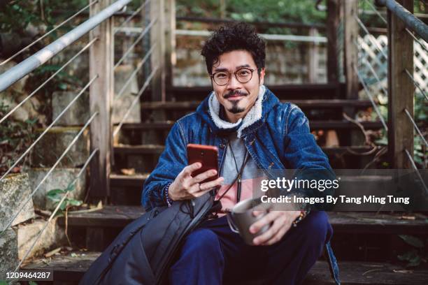 young asian man smiling joyfully at camera while using smartphone in country side - accesorio para ojos fotografías e imágenes de stock