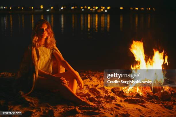 young caucasian woman with a plaid on her shoulders sitting near a fire at night on the background of the river. - bonfire beach stock pictures, royalty-free photos & images