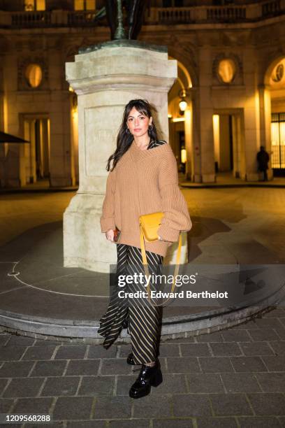 Guest wears a pale brown v-neck oversized wool pullover, a yellow bag, a striped skirt, black leather shoes, earrings, outside the MyTheresa x Amina...