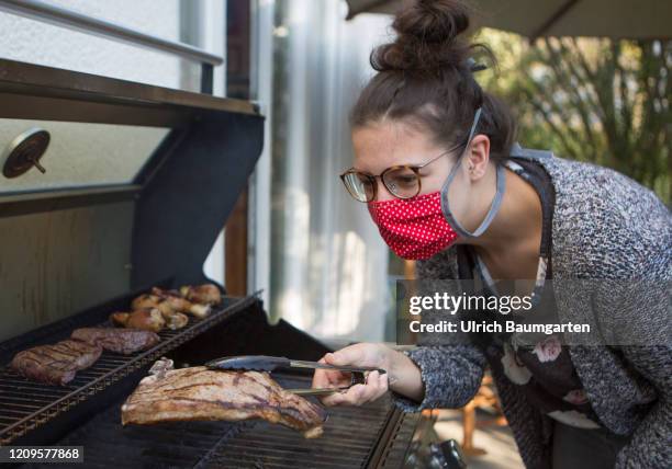 The corona virus keeps the world in suspense! Young woman with protective mask grills a steak. Symbol photo on the topics COVID-19, SARS-CoV-2,...