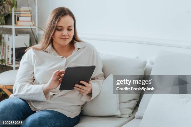 business at home: plus size caucasian woman sitting on a sofa using a tablet - full figure stock pictures, royalty-free photos & images