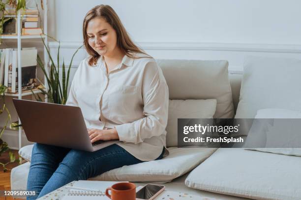 negocios casuales: mujer feliz más tamaño sentada en su sala de estar y trabajando en un ordenador portátil - curvy woman fotografías e imágenes de stock