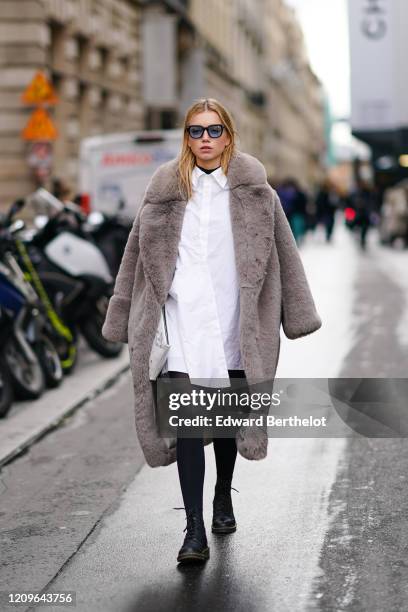 Guest wears sunglasses, a white shirt, black opaque tights, black army-style boots, a white bag, a fluffy taupe-color faux-fur coat, during Paris...