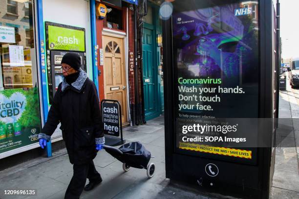 Woman wearing a face mask walks past a Coronavirus public information campaign poster saying 'Wash your hands. Don't touch your face' in London.