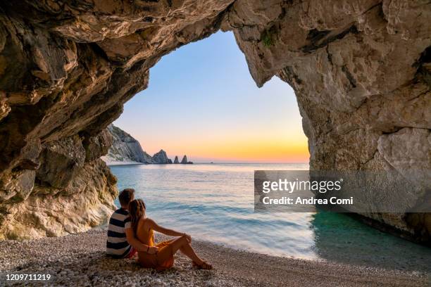 young couple enjoying the sunrise from a cave at two sisters beach (le due sorelle). conero, italy - adria tour stock-fotos und bilder