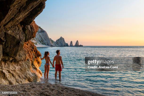 young couple enjoying the sunrise at two sisters beach (le due sorelle). conero, italy - ardia stock-fotos und bilder