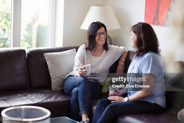 two happy women sitting together on a sofa with cups of coffee - friends talking living room stock pictures, royalty-free photos & images