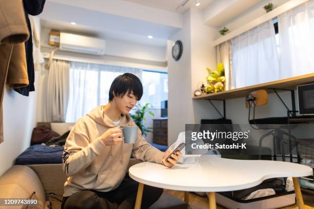 若いハンサムな男の子がコーヒーを飲んで、自宅でスマートフォンを使用して - 美大生　日本 ストックフォトと画像