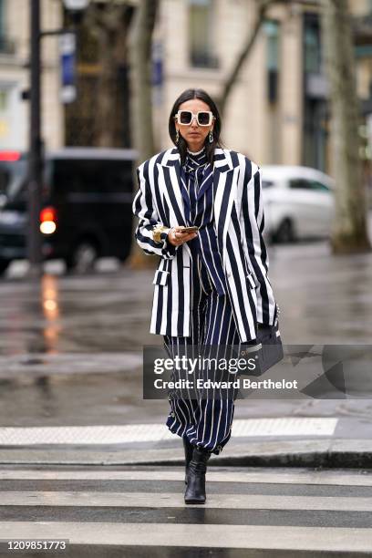 Gabriella Berdugo wears white sunglasses, earrings, a bracelet, rings, a navy-blue jacket with white stripes, matched pants with side-buttons, a...