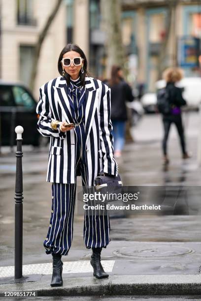 Gabriella Berdugo wears white sunglasses, earrings, a bracelet, rings, a navy-blue jacket with white stripes, matched pants with side-buttons, a...