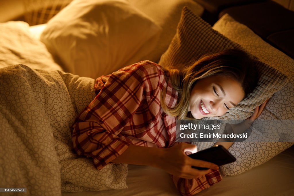 Beautiful young woman using mobile phone in bed at night