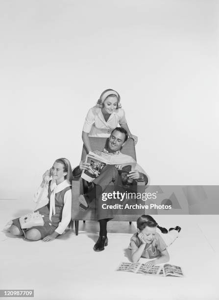 Woman standing behind man sitting on chair reading magazine, children playing around