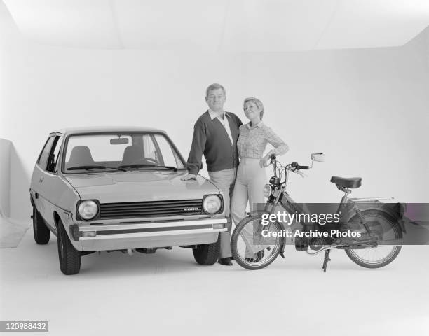 Mature couple standing beside Ford Fiesta and Puch moped, smiling, portrait