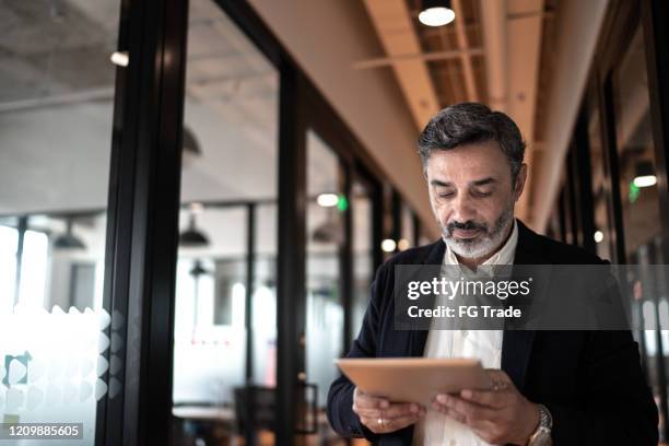 mature businessman walking and using digital tablet at corridor office - investment research stock pictures, royalty-free photos & images