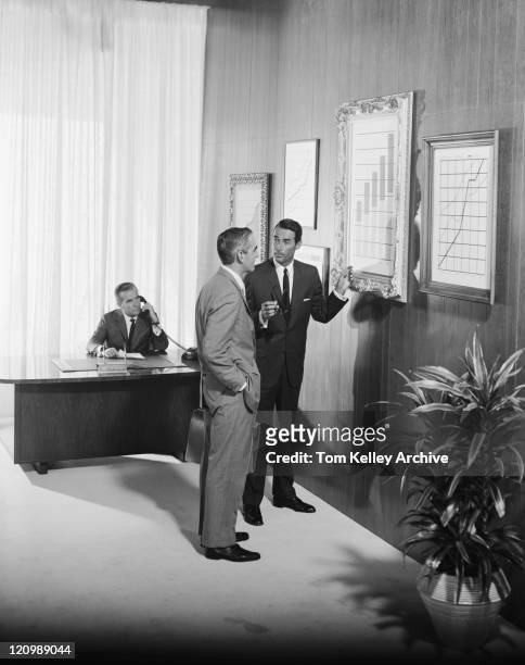 two businessman discussing at bar chart while another man using telephone in background - archival man stock pictures, royalty-free photos & images
