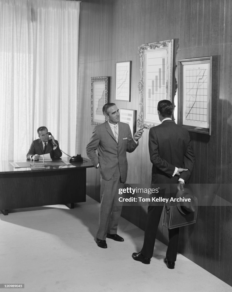 Two businessman discussing at bar chart while another man using telephone in background