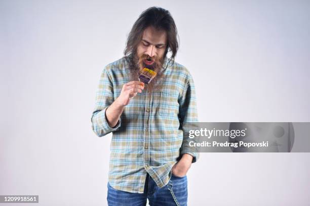 portrait of young man artist trying to byte paint brush on studio background - kuriose mode stock-fotos und bilder