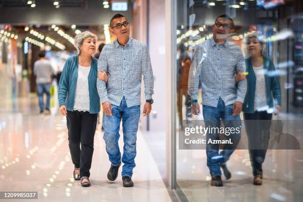 chinese senior shoppers walking arm in arm at mall - couple shopping in shopping mall stock pictures, royalty-free photos & images