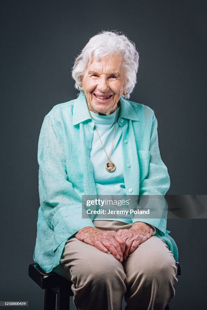 Sweet, Joyful, Happy, Wise Elderly Senior Caucasian Woman Smiling, Wearing Her Favorite Teal Blue Shirt and Sitting on a Stool Against a Gray Background in Studio with Copy Space