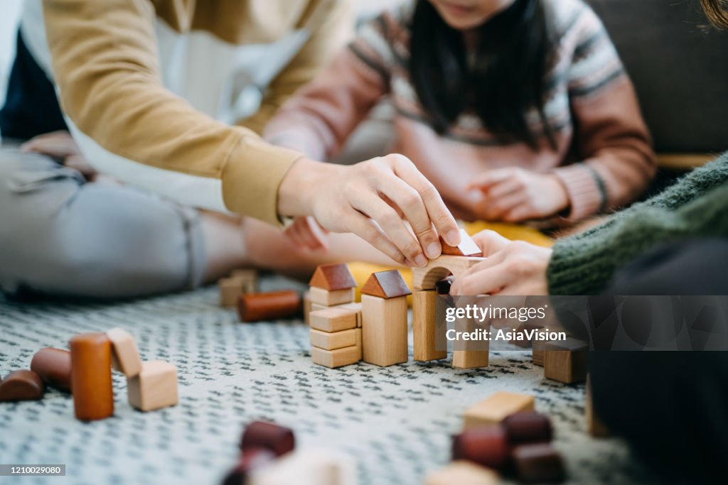 Close up of joyful Asian parents sitting on the floor in the living room having fun and playing wooden building blocks with daughter together