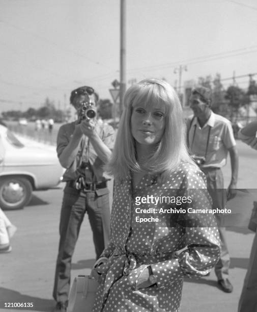 French actress Catherine Deneuve, wearing a polka-dotted dress and holding a handbag, portrayed while standing between cameramen and photographers,...