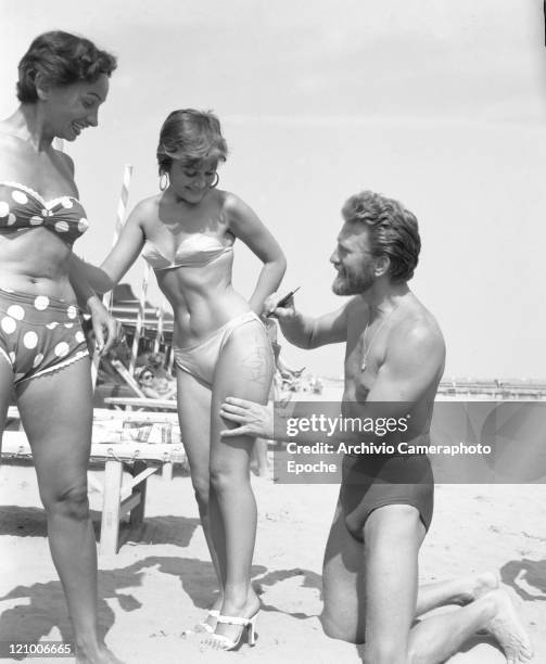 American actor Kirk Douglas, wearing a swimming suit and a necklace chainlet, signing on the thigh of the paintress Novella Parigini, wearing a...