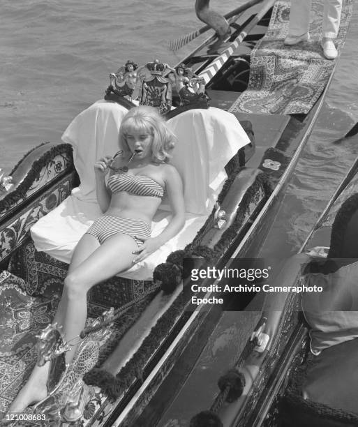 American actress Sue Lyon, earing a strapless, striped bikini and holding Lolita sunglasses, portrayed while lying on a gondola, Venice, 1962.
