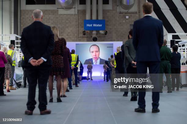 People observe social distancing measures as they stand amongst Hospital cubicles, as a giant screen displays an image of Britain's Health Secretary...