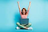 Full size photo of excited ginger hair woman sit floor legs crossed work laptop finish start-up report scream yeah raise fists wear green white sweater footwear isolated blue color background