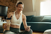 Happy athletic woman using mobile phone while drinking smoothie at home.
