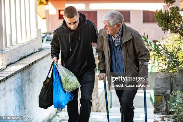 giovane uomo adulto che aiuta l'uomo adulto anziano con le stampelle a camminare e portare la spesa - bidello foto e immagini stock