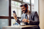 Young smiling bearded businessman sitting in coffee shop, reading something funny on smart phone and stirring coffee.