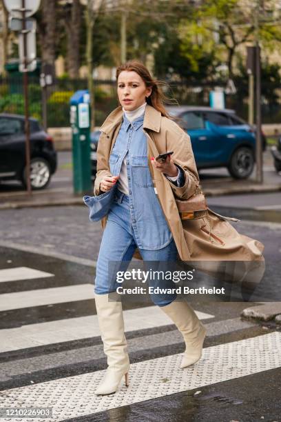 Guest wears earrings, a white turtleneck, a blue denim shirt, blue jeans, a sand-color trench coat, white pointy heeled knee-high boots, a Fendi bag,...