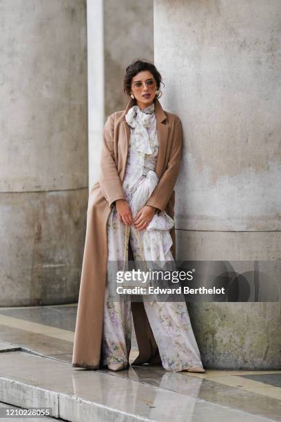 Ketevan Giorgadze wears sunglasses, earrings, a camel long coat, a white flowing jumpsuit with colorful floral prints, a lavaliere and a belt bow,...