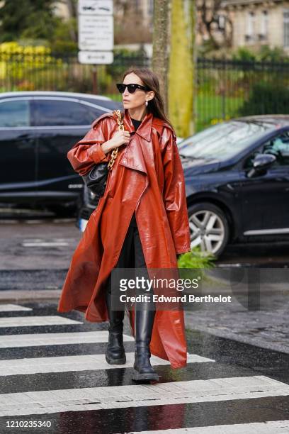 Chloe Harrouche wears sunglasses, earrings, a Bottega Veneta bag with a golden chain handle, a red leather trench long coat, black pants, black...