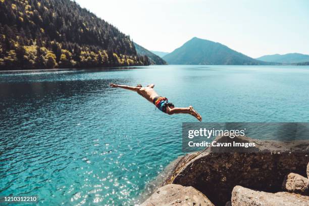 side view of young male jumping in alpine lake in front of mountains - salto desde acantilado fotografías e imágenes de stock