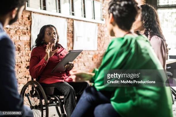 businesswoman in wheelchair leading group discussion in creative office - coinvolgimento dei dipendenti foto e immagini stock