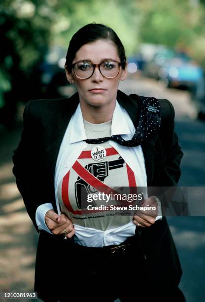 Portrait of Canadian-American actress Margot Kidder , dressed in a suit and tie, as she pulls open her shirt to reveal a t-shirt that reads 'National...