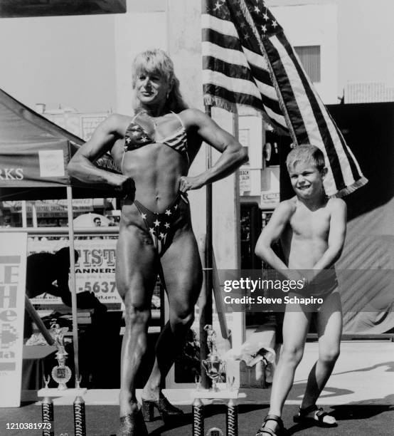 View of a pair of contestants, a woman in a 'stars and stripes' bikini and a young boy, both posing, during a Muscle Beach Venice bodybuilding...