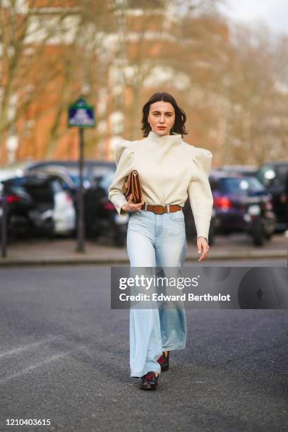 Mary Leest wears a white top with shoulder pads, a brown leather clutch, a brown leather belt, pale blue denim flared pants, shoes, outside Lacoste,...