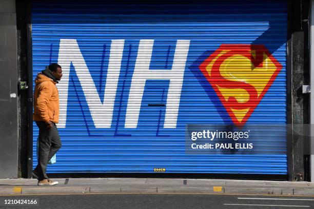 Pedestrian walks past graffiti depicting the logo of Britain's NHS , merged with the emblem or badge of the fictional super heros Superman and...