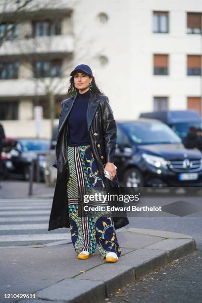 Julia Haghjoo wears a blue cap, a long black leather coat, a navy blue pullover, Lacoste flared pants with printed geometric patterns, earrings,...