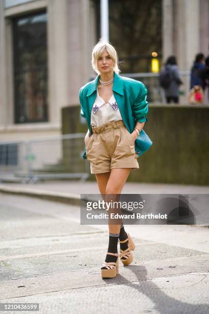 Xenia Adonts wears a green leather jacket with shoulder pads, a low neck shirt with printed flowers on the collar, pale brown shorts, black socks,...