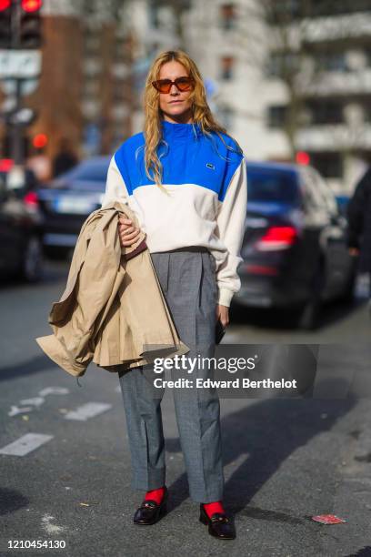 Blanca Miro wears sunglasses, a blue and white Lacoste jacket, gray pants, red socks, black leather shoes, holds a trench coat, outside Lacoste,...