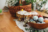 Stylish easter eggs natural dyed on background of homemade easter cake, candle, green branches and flowers with wicker basket on wooden rustic table. Easter Food for sanctify