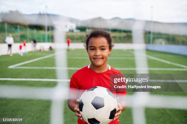 a small girl with soccer ball standing outdoors on football pitch, looking at camera. - jugendfußball stock-fotos und bilder