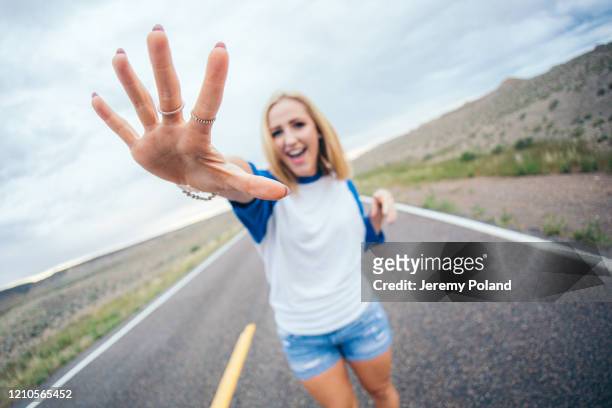 fisheye wide angle shot of young beautiful blonde caucasian 20-year-old adult woman holding up 5 fingers open hand palm while standing in the street country road in the colorado desert on a cloudy overcast day - short hair photos et images de collection