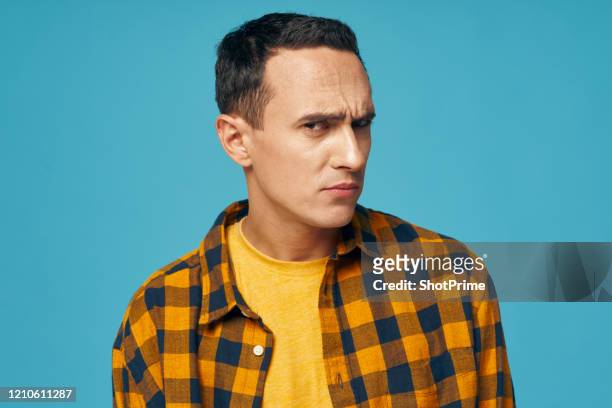 skeptical young man looks with suspicion, blue background, yellow shirt - angry faces stock-fotos und bilder