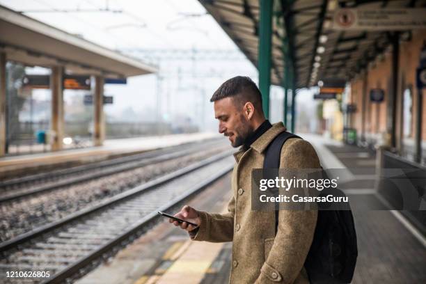 handsome turist titta på innehåll på sin mobiltelefon väntar på tåg att komma - stock photo - train platform bildbanksfoton och bilder