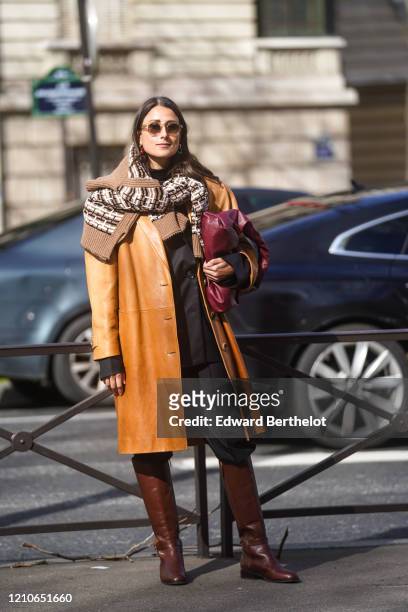 Julia Haghjoo wears sunglasses, a brown leather coat, a purple leather bag, a wool pullover with printed patterns, black pants, leather boots,...
